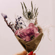 Hand holding a small bouquet of pink flowers and dried elements against a light background